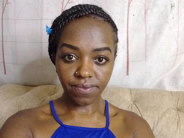 CrystalEbonyQueen with dark skin, braided hair, blue hair clip, nose ring, blue halter top, subtle smile, sitting on beige couch, looking at camera.