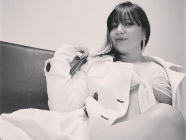 RoxanneBlair with shoulder-length dark hair, wearing a white button-up coat, exposing her small tits, sitting on a leather chair, holding her hair, smiling slightly, black-and-white photo.