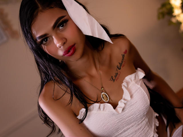 CrystalDavids with dark hair, white dress, red lipstick, and a white paper bow, has a tattoo on her chest and wears a necklace. Boobs, cute, and looking at camera.