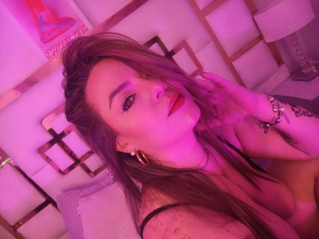 AvaMia with pink-tinged hair, red lipstick, and gold earrings, leaning forward, showing cleavage, touching her neck, wearing a bracelet, in a purple-lit room.