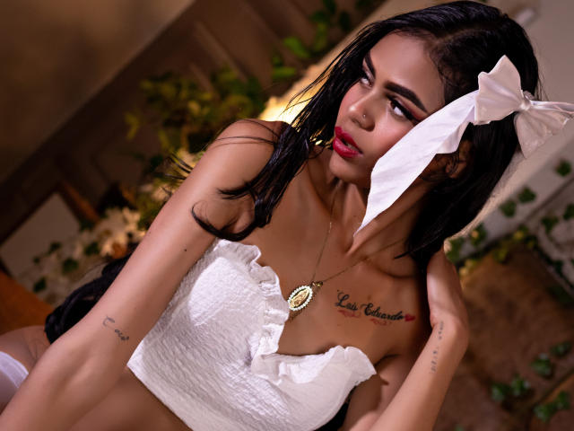 CrystalDavids with dark hair, white bow, and strapless white top, showing her tits and tattoos, wearing a necklace, looking to the side with red lipstick, in a bedroom.