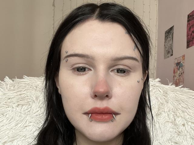 Yanira Stroupe tem pele clara, cabelo preto, olhos verdes, um piercing no nariz e dois piercings faciais com um anel no lábio inferior. Ela está olhando para a câmera com uma expressão neutra..