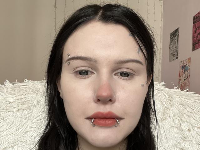 Yanira Stroupe ha la pelle chiara, i capelli neri, gli occhi verdi e diversi piercing sul viso: sul naso, sul labbro inferiore e sopra le sopracciglia. È seduta su un cuscino bianco e soffice..