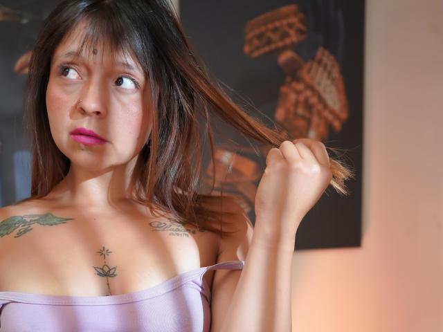 QueenFit, con capelli castani, rossetto rosa e tatuaggi, che tiene una ciocca di capelli, indossa una maglietta viola con una spalla scoperta, mostrando i suoi piccoli seni. Illuminazione soffusa, leggero rossore sulle guance..