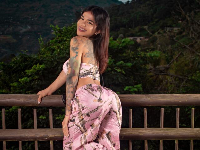 PrincessMelim with tattoos, smiling, wearing pink floral bra and pants, leaning on wooden railing, showing her ass, brown hair, dark skin, looking back, outdoor setting.