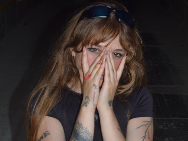UwuMaddiee with hands covering her face, brown wavy hair, black t-shirt, tattoos on fingers and arms, blue sunglasses on head, red and yellow nail polish, slightly teary eyes.
