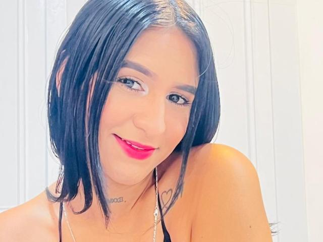 Lilii Adams, con i capelli neri lucidi, il rossetto rosso e un tatuaggio sotto il mento, sorride alla fotocamera, indossando un top nero con spalline e una collana d'argento..