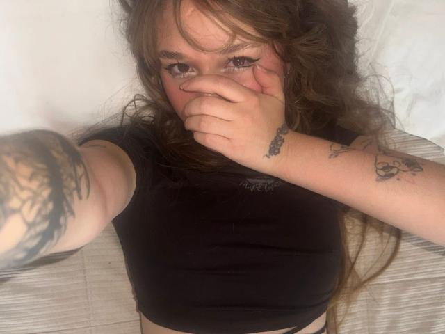 UwuMaddiee with long, wavy brown hair, covering her mouth with her hand, wearing a black top, showing tattoos on her right arm, lying on a bed.