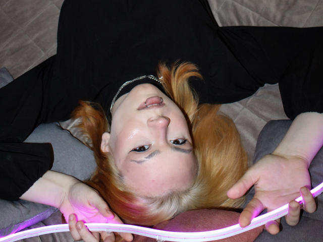 Remy Calhoun, con cabello rojo, camisa negra, acostado, sosteniendo un cable neón rosa, mirando hacia arriba, labios ligeramente entreabiertos, piel pálida, maquillaje sutil, piercing visible en la nariz, relajado..