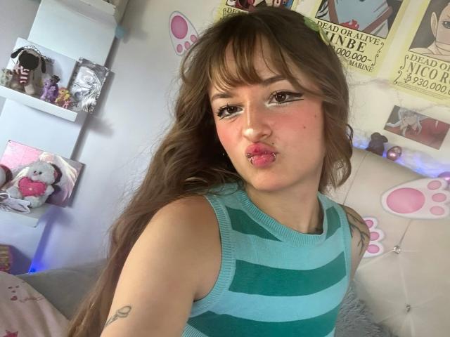 UwuMaddiee, con cabello castaño largo, labios rosados y delineador de ojos marcado, vestida con una camiseta de tirantes a rayas verdes y azules, mostrando un pequeño tatuaje en su brazo..