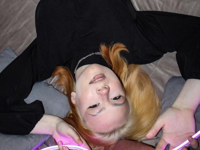 RemyCalhoun with blonde hair, black top, and black eye mask, lying on gray and brown pillows, tongue out, holding pink neon lights, arms above head.