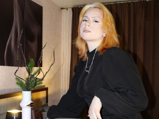 RemyCalhoun with red hair, black dress, and silver necklace, looking slightly up, has pale skin and small tits, sitting in a room with a brown curtain and vase.