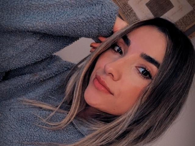 AdharaCooperr with blonde highlights, fluffy gray sweater, raised arm, and subtle smile, looking directly at camera. Her eyebrows are thick, and she has full, pink lips.