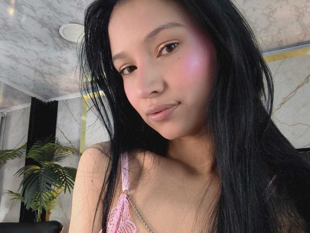 SophieDelaney with long black hair, pink blush, and a subtle smile, wearing a pink lace top, showing her small tits and smooth, fair skin.