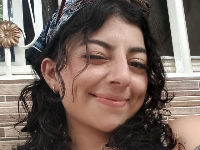 AnntonellaWatson has curly black hair, a denim headscarf, and is smiling with eyes half-closed, showing her nose and slightly parted pink lips. She wears small gold earrings.