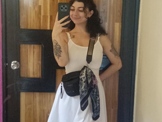 AnntonellaWatson with curly brown hair, white tank top, white skirt, black bag, and striped necktie, holding phone, tattoos on arm, smiling, standing in front of wooden door.