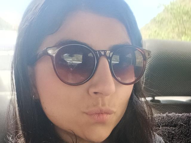 AnntonellaWatson with dark brown tortoiseshell sunglasses, pouty lips, and black straight hair, sitting in a car, reflecting a vehicle outside. Her ass is not visible.