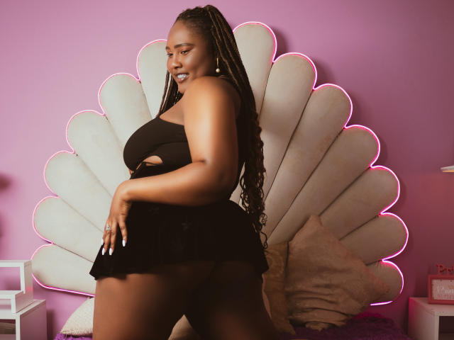 SkylerNaomi with dark skin, long braids, black cut-out dress, showing her ass, standing in front of a white fan-shaped wall with pink neon trim. She's looking over her shoulder.