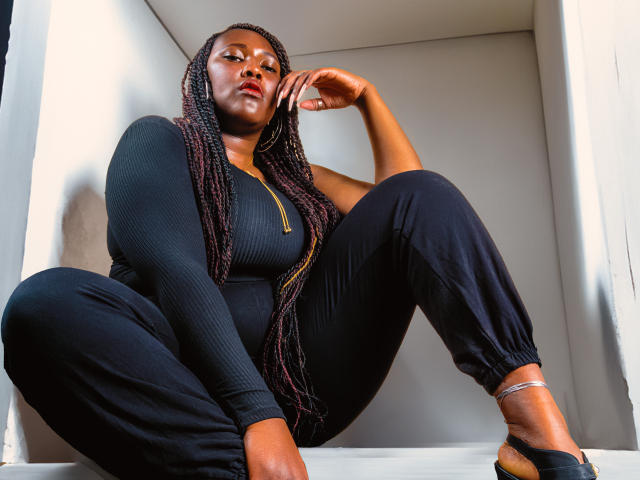 SkylerNaomi with dark skin, long braided hair, black ribbed bodysuit, and black pants, sitting with legs spread, hand on cheek, looking down, wearing black sandals.
