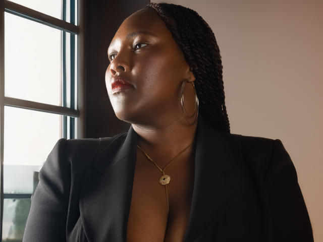 SkylerNaomi with dark skin, braided hair, large hoop earrings, gold necklace, black blazer, and subtle cleavage, looking to the side, near a window.
