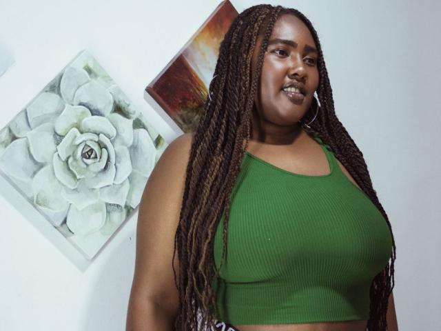 SkylerNaomi with dark brown skin, long braids, green ribbed crop top, showing her big boobs, standing, smiling, wearing large hoop earrings, in a simple indoor setting.