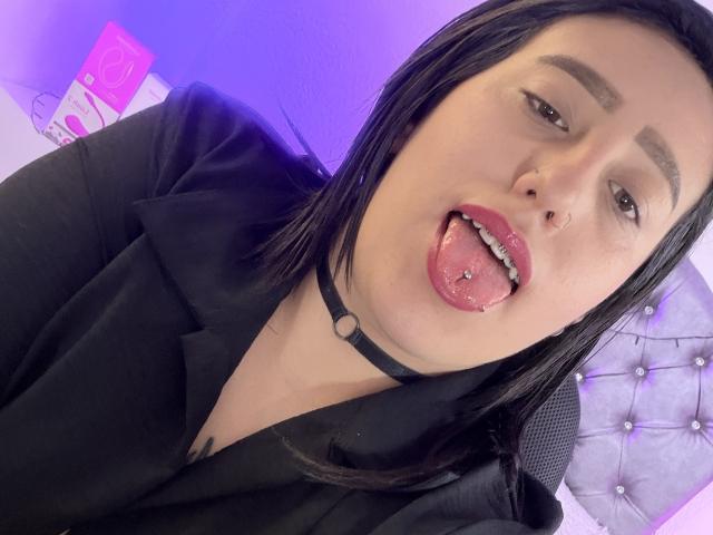 NatalieHarris with black hair, black choker, black shirt, tongue out, tongue piercing, nose piercing, and visible nipple tattoo, in a selfie. Purple light in the background.