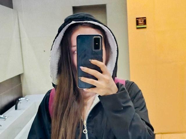 VioletaNova mit langen braunen Haaren, schwarzer Kapuzenjacke, rosa Rucksack, hält ein Handy vor ihrem Gesicht, Spiegelbild in einem Badezimmer. Ihr Handy verdeckt ihre Augen..