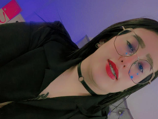 NatalieHarris with glasses, black choker, red lipstick, blue face markings, black jacket, black hair, blue-lit background, nose piercing, tattoo on chest.