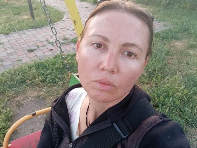 ZoyaPask has a serious face, brown eyes, and is wearing a black vest with straps, standing on a playground, grass, and brick path in the background.