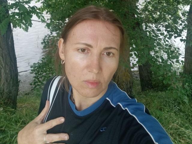 Zoya Pask, met volle rode lippen, bruine ogen en een serieuze uitdrukking, draagt een zwarte en blauwe sportshirt en houdt haar shirt met één hand vast, onder een groene boom..