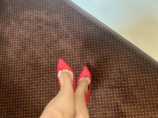 MoonLiza's legs with red pointed-toe heels, showing her bare toes and feet on a brown, patterned carpet. Her skin is light, and her feet are slightly crossed.