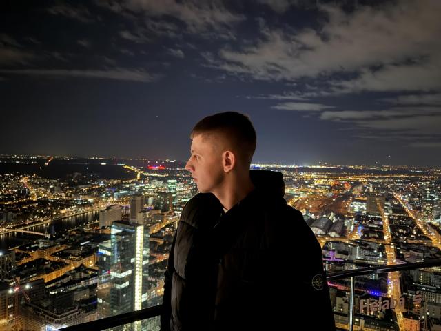 CrystalAlex, con un taglio di capelli corto e indossando un giubbotto nero imbottito, è in piedi su un balcone di notte, osservando una città illuminata con un cielo nuvoloso..