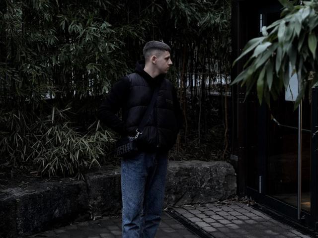 CrystAlex está em pé, vestindo um colete acolchoado escuro, jeans azuis e luvas pretas, com cabelo curto e uma expressão séria, ao lado de bambu e uma porta de vidro..