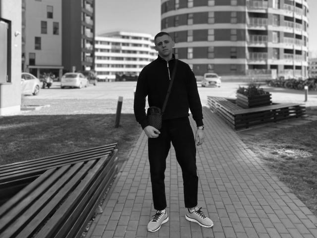Schwarz-weißes Foto von CrystalAlex, die auf einem Ziegelweg steht. Sie trägt eine schwarze Jacke, eine Hose, Turnschuhe und eine Umhängetasche. Sie ist kahlköpfig, muskulös und hat einen ernsten Gesichtsausdruck. Im Hintergrund sind Gebäude und Autos zu sehen..