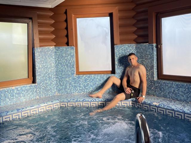 CrystalAlex, sem camisa, usando shorts de banho pretos, sentado em uma banheira de hidromassagem com azulejos azuis, perna direita na água, perna esquerda na borda, paredes com painéis de madeira, janelas, luz da manhã, jatos de água, relaxado, atraente..