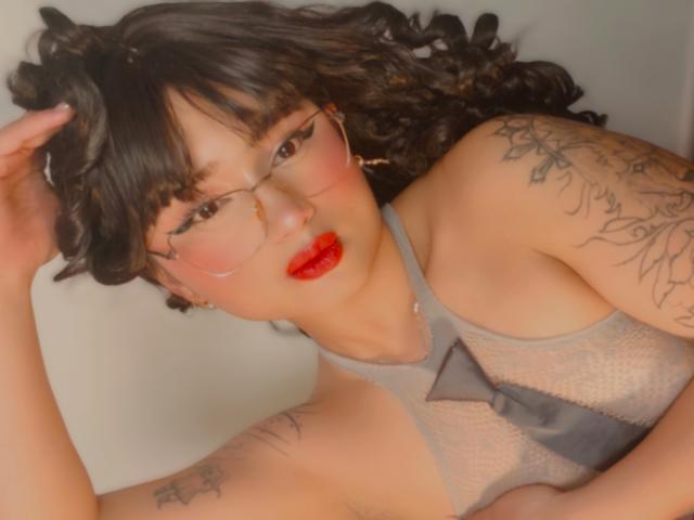 AnnieGrey with curly black hair, glasses, red lipstick, tattoos on arms, wearing a gray mesh top with a black bow, lying down, hand in hair. Boobs, tits, pussy, ass.