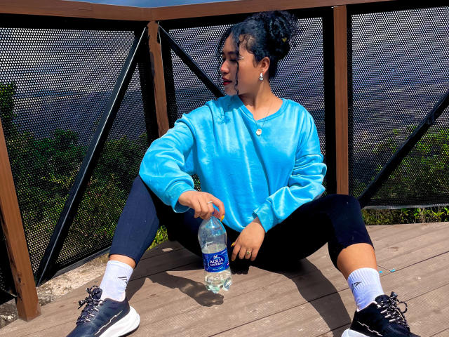 SophieSonyas with black curly hair, blue sweatshirt, black capri pants, white socks, black sneakers, holding a water bottle, sitting on wooden deck, black mesh fence in background.