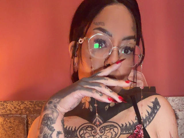 MistressSinful with glasses, red nails, and tattoos, touching her lips with a finger. She has a nose ring and a green light reflection in her eye. Red wall background.