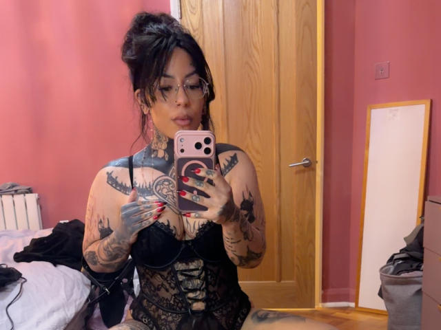 MistressSinful, con tatuaggi, occhiali e lingerie in pizzo nero, tiene in mano un telefono rosa, scatta un selfie allo specchio in una stanza rosa, mostrando i suoi seni e i tatuaggi..