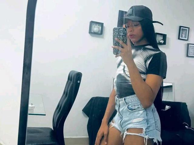 NashlaLonga with long black hair, wearing a black cap, black and white t-shirt, and ripped blue denim shorts, taking a mirror selfie in a room. Her ass is visible.