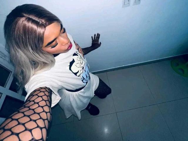 NashlaLonga, con capelli biondi e sfumati, indossa una t-shirt bianca con una grafica, maniche a rete nere e guanti, e fa una smorfia in un selfie, in piedi su un pavimento piastrellato..