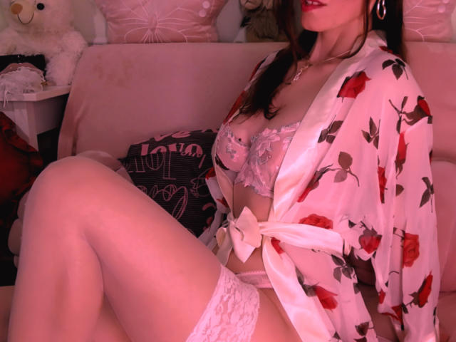 BlueNightX wearing a floral silk robe, exposing her tits in a lace bra, white thigh-high stockings, and red lipstick, sitting on a pink sofa with a teddy bear.