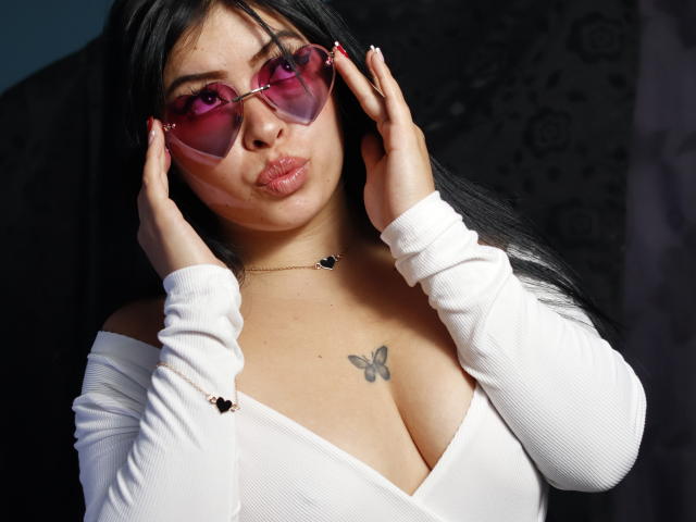 KateWelsin with pink glasses, pouty lips, white off-shoulder top, butterfly tattoo on chest, holding glasses with hands, black hair, tan skin, looking at camera. Boobs visible.