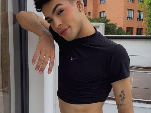 Aresxx leans against a wall, wearing a black Nike crop top, showing a rose tattoo on his right arm, with a slight smile, short curly hair, and pink lipstick.