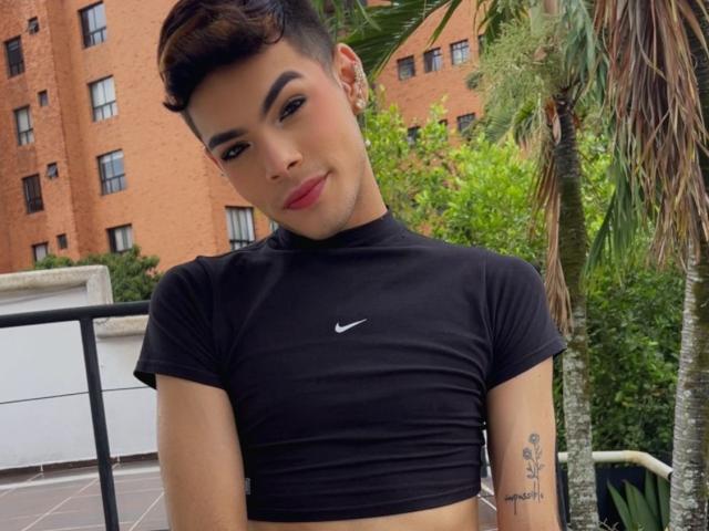 Aresxx, com cabelo castanho curto, top cropped preto da Nike, seios pequenos, tatuagem no braço direito, batom vermelho e piercing na orelha, está em pé do lado de fora, com vegetação e uma palmeira ao fundo..