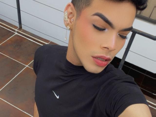 Aresxx, avec ses cheveux courts et foncés, ses sourcils épais et ses lèvres roses, porte un t-shirt noir Nike, a une boucle d'oreille en perle et un anneau en or, les yeux fermés, et regarde vers le bas..