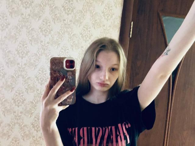 MelbaCaffee, with pale skin and blonde hair, takes a selfie in a mirror, wearing a black "PINK" shirt, raising her right arm, showing a small tattoo.
