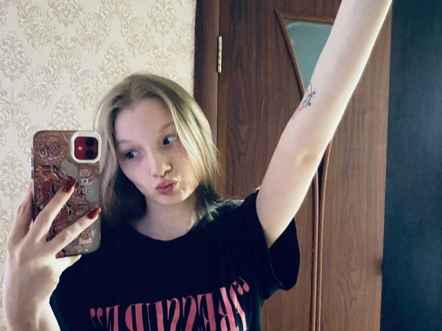 MelbaCaffee, con cabello rubio, labios carnosos, levantando el brazo derecho, mostrando un tatuaje, sosteniendo un teléfono con una funda roja, vistiendo una camiseta negra con la palabra "Supercross", en un selfie frente al espejo..