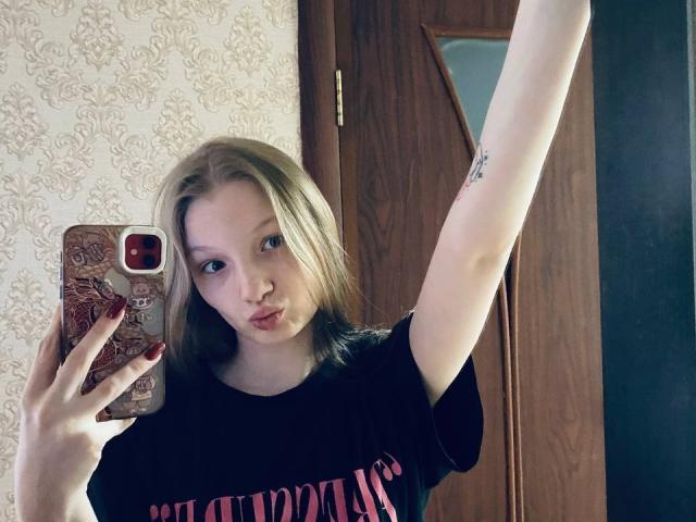 MelbaCaffee mit blonden Haaren, die die Lippen zusammenpresst, in einem schwarzen T-Shirt, hält ein Handy mit roter Hülle, zeigt ein Tattoo auf dem erhobenen Arm, im Spiegel-Selfie, mit einer Holztür im Hintergrund..