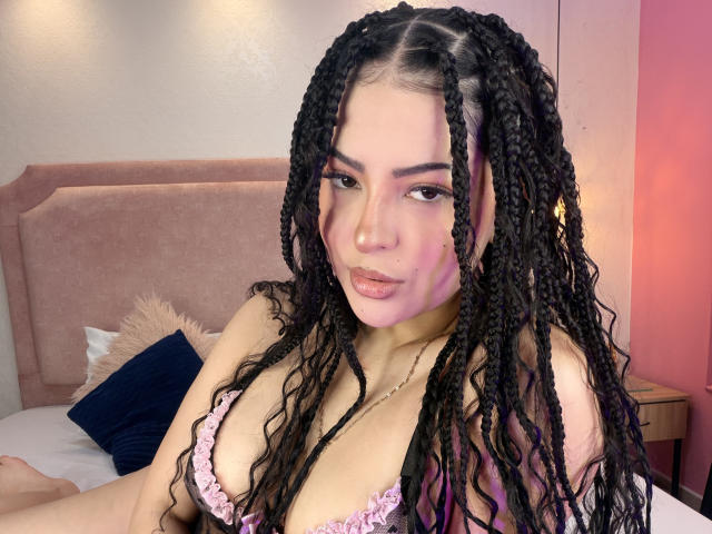 RheaVolk with long black braided hair, pink eyeliner, and a pink frilled bra, showing her tits, sitting on a bed with a pink headboard. She's looking at the camera.